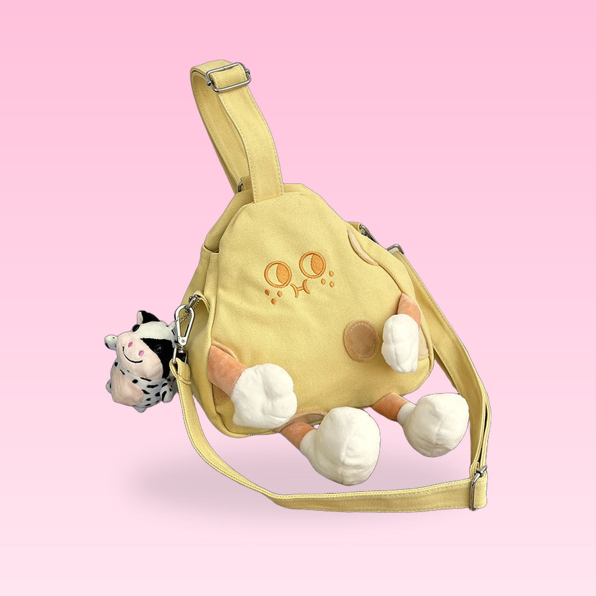 Yellow canvas cheese wedge crossbody bag with an embroidered face, plush legs and a cow charm on a pink background