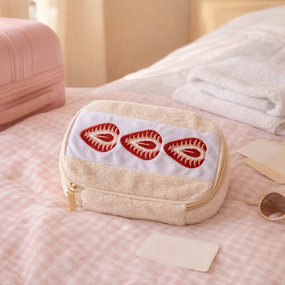 Strawberry sando makeup bag on a pink gingham bed beside a pink suitcase, packed for a trip