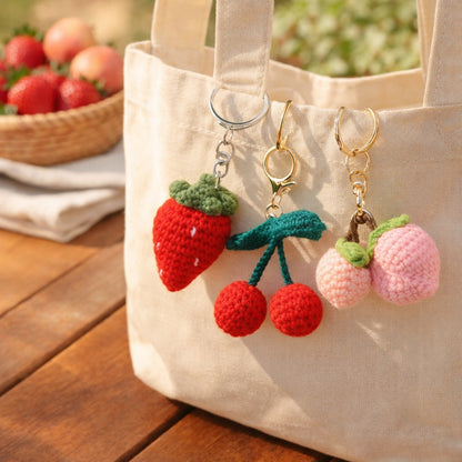 Crochet strawberry, cherry and peach keychains clipped to a canvas tote bag at a farmers market table