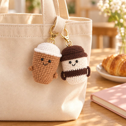 Latte and Mocha crochet coffee cup charms clipped to a cream canvas tote on a cafe table with a croissant
