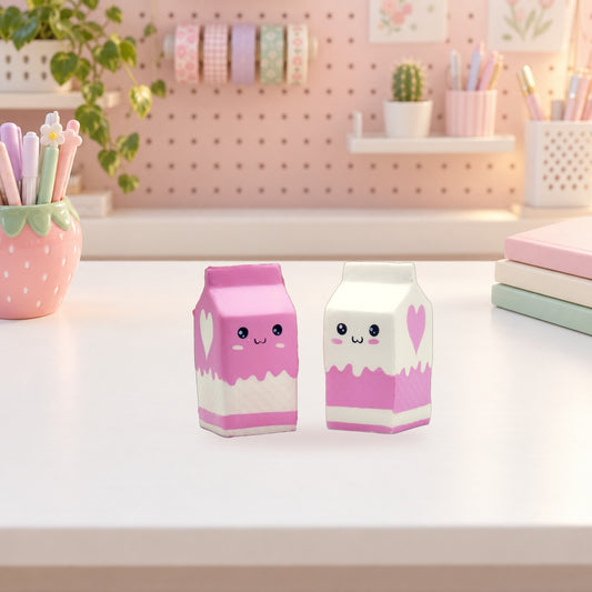 Two milk carton squishies on a pastel desk beside a strawberry pen pot and a stack of notebooks