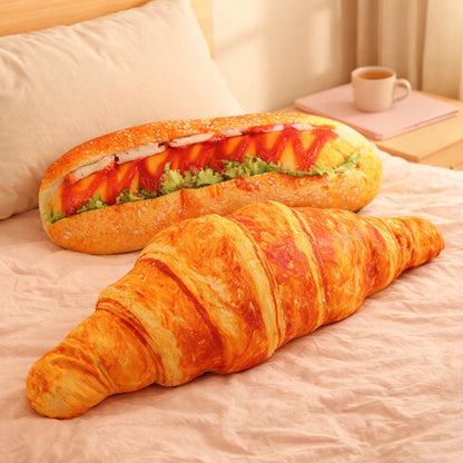 Croissant and hot dog hug pillows lying across a bed with a pink duvet, a cup of tea in the background