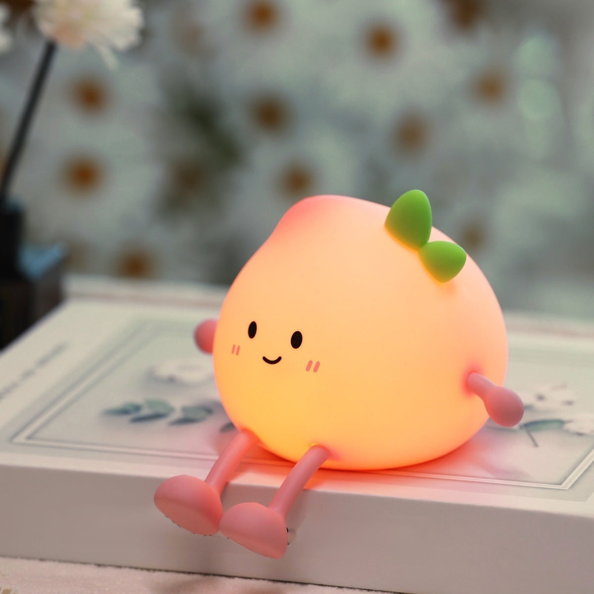 Peach night light glowing warm while perched on the edge of a book with its legs dangling