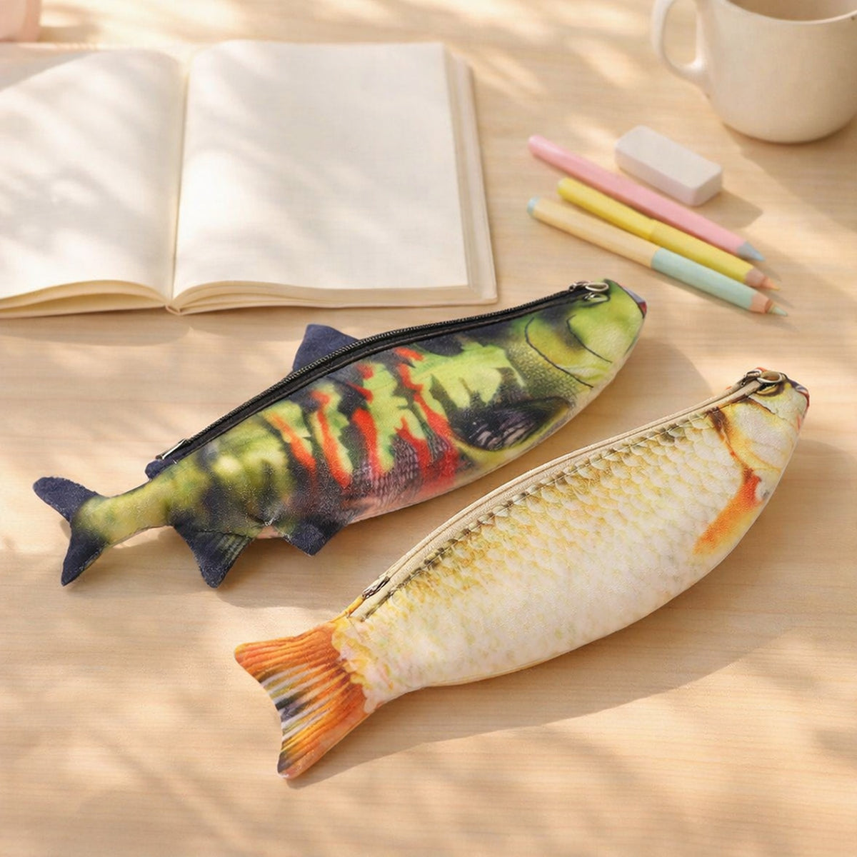 Two fish pencil cases, a sockeye salmon and a golden crucian carp, on a sunny study desk with a notebook and pastel pencils