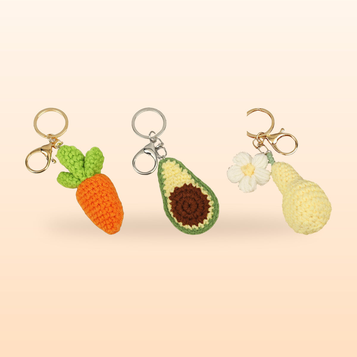 Three crochet keychains, a carrot, an avocado and a pear with a flower, on a soft peach background