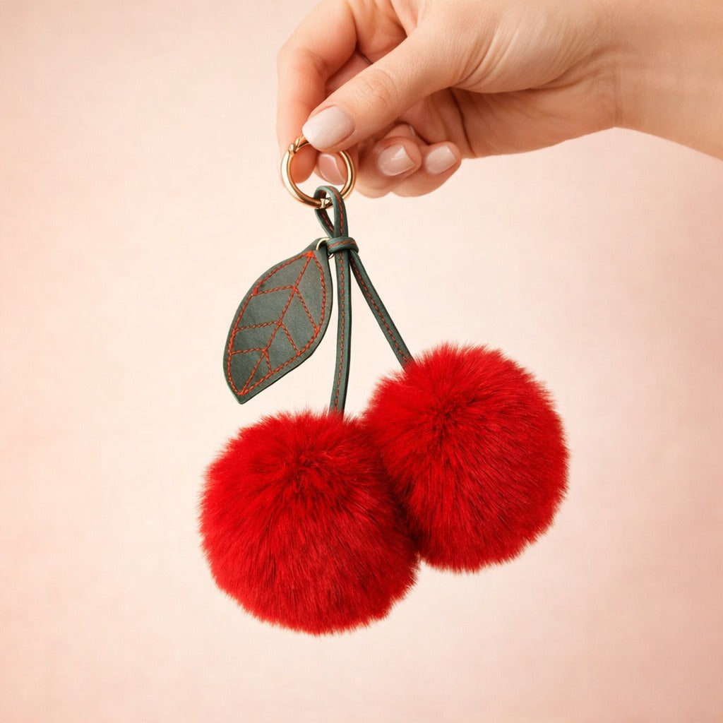 Hand holding the red cherry pom-pom bag charm by its gold ring to show its size