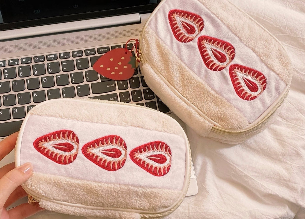Strawberry sando makeup bags next to a laptop and held in a hand to show their real size