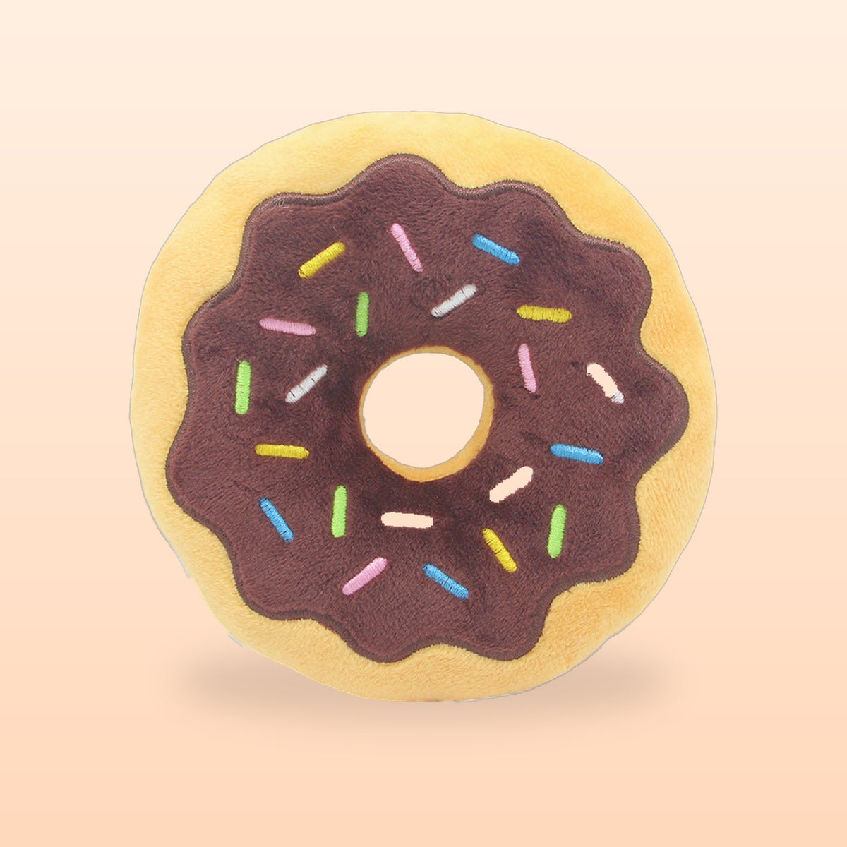 Chocolate donut squeaky plush pet toy with embroidered sprinkles, on a soft peach background