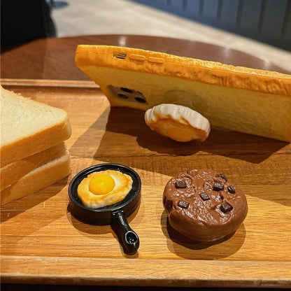 Toast bread iPhone case standing on a wooden tray beside fried egg, pan and cookie grips