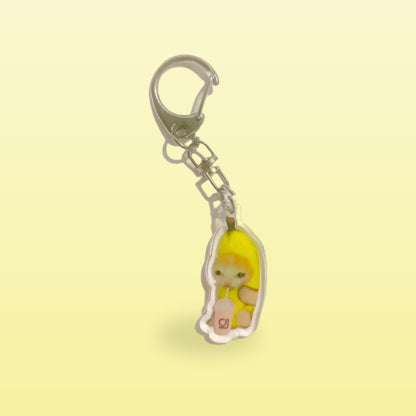 Banana cat meme acrylic keychain holding a pink drink with a silver clip, on a butter yellow background