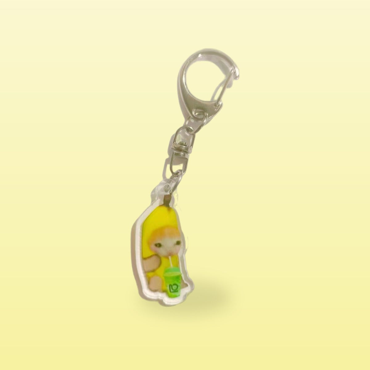 Banana cat meme acrylic keychain holding a green drink with a silver clip, on a butter yellow background