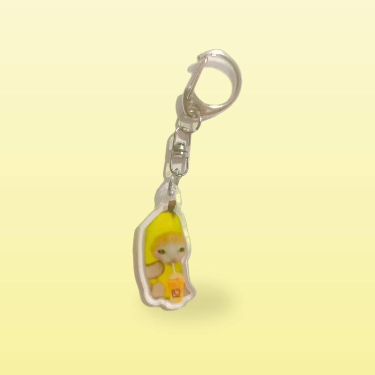 Banana cat meme acrylic keychain holding an orange drink with a silver clip, on a butter yellow background