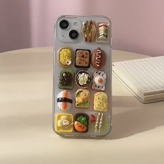 Sushi Symphony clear iPhone case with tiny resin sushi and toast charms, turned in a hand on a pastel desk