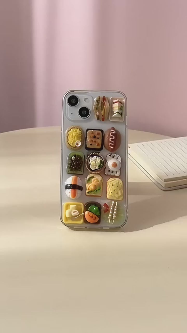 Sushi Symphony clear iPhone case with tiny resin sushi and toast charms, turned in a hand on a pastel desk