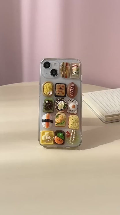 Sushi Symphony clear iPhone case with tiny resin sushi and toast charms, turned in a hand on a pastel desk