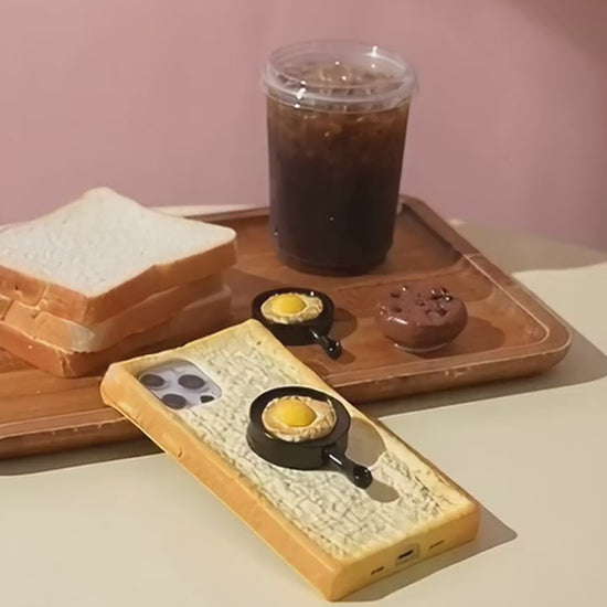 Toast bread iPhone case with the frying pan grip, picked up and turned in a hand on a pastel breakfast table
