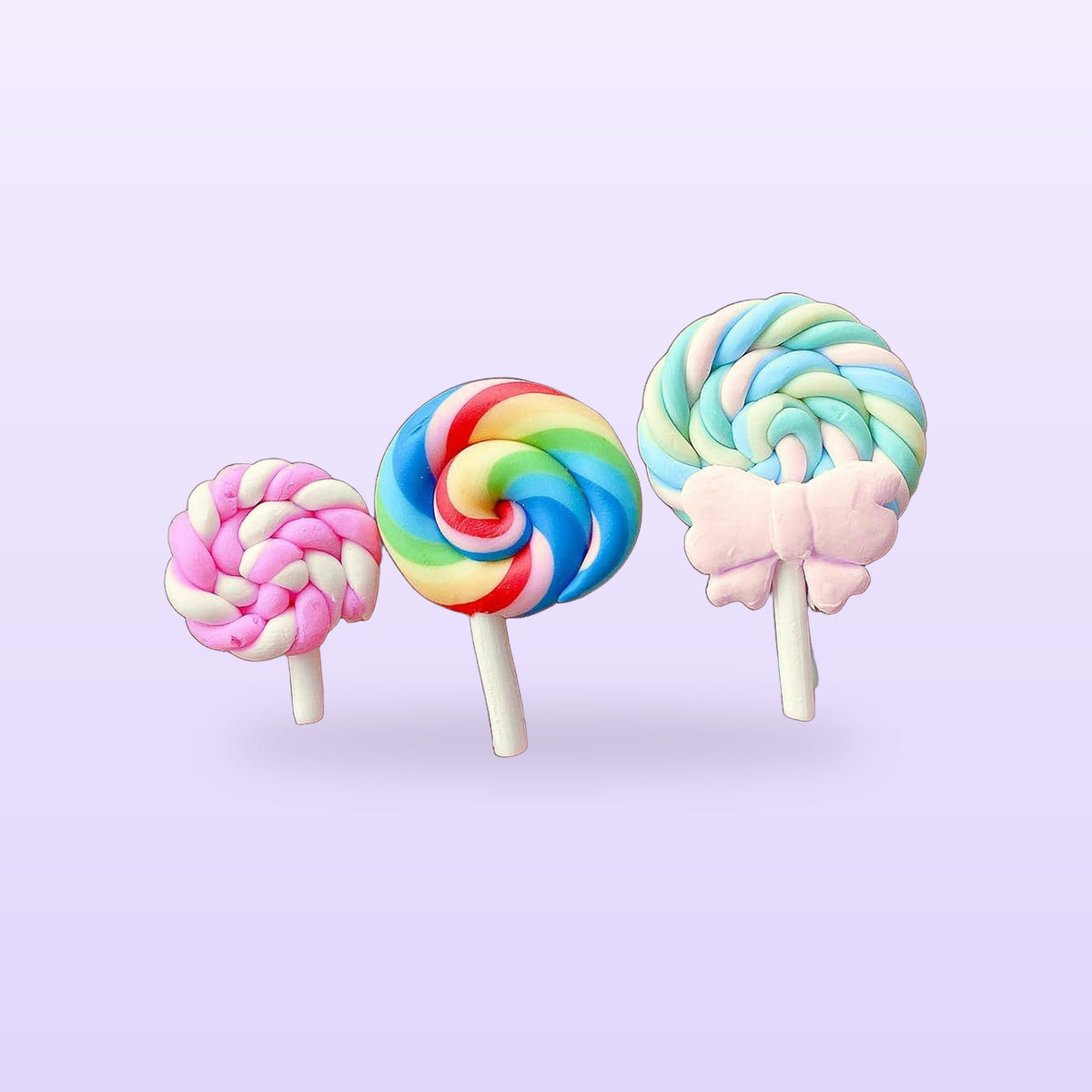 Three candy resin charms, pink, rainbow and pastel blue swirl lollipops, on a lilac background