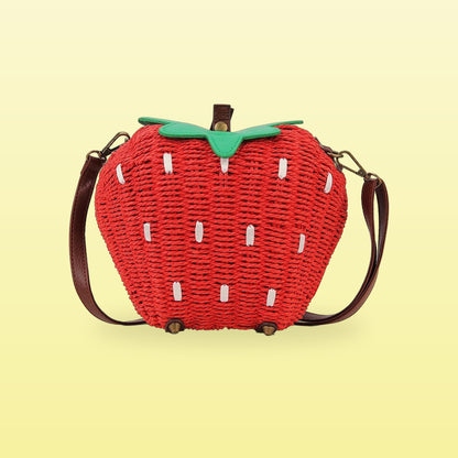 Red woven straw strawberry crossbody bag with a green leaf top and brown strap on a butter yellow background