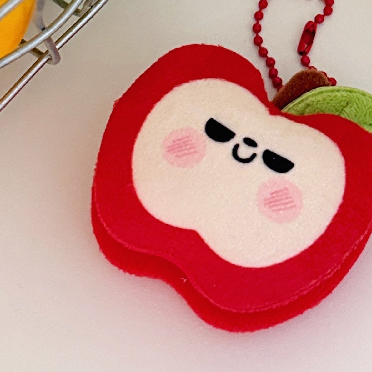 One red plush apple keychain with white face, black sleepy eyes, pink cheeks, green leaf and red bead chain.